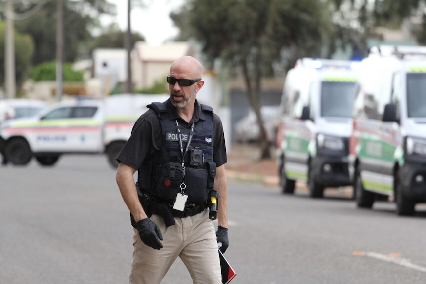 A police officer wearing a vest walks down the street, with ambulances and a car in the background.