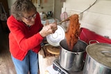 A woman uses a wooden spoon to pull a ball of dark orange wool out of a pot of hot water on an outdoor stove.