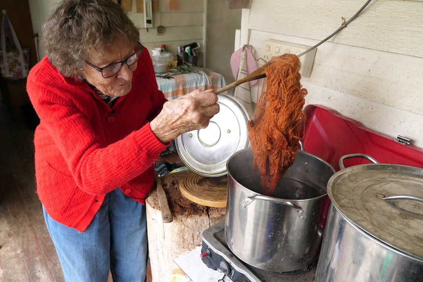 Eileen uses a wooden spoon to pull a ball of dark orange wool out of a pot of hot water on an outdoor stove.