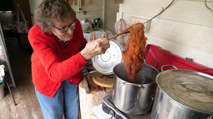 A woman uses a wooden spoon to pull a ball of dark orange wool out of a pot of hot water on an outdoor stove.