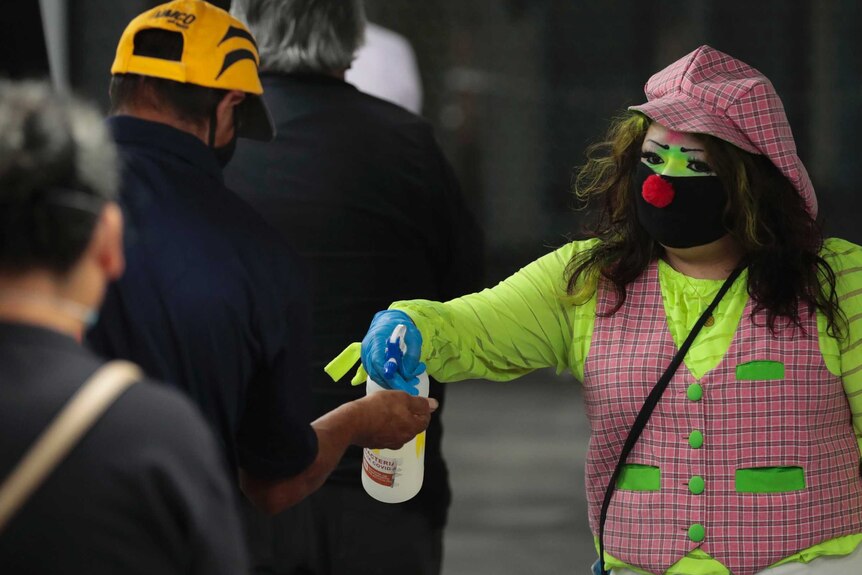 A woman with a black face mask, red nose and clown make up, uses a spray bottle to squirt liquid into a man's hand
