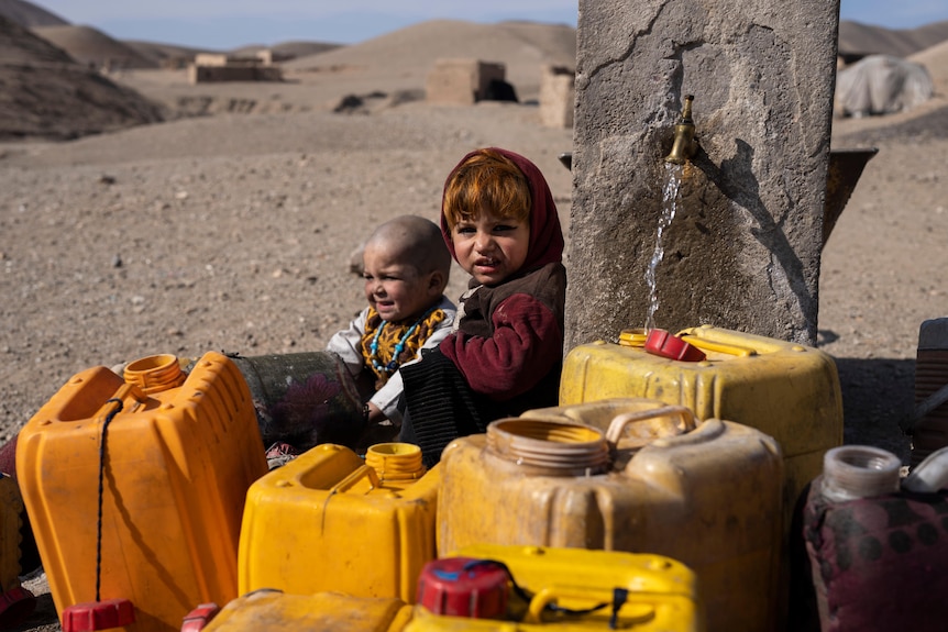 Two Afghan children sit next to canisters and a spigot running water into a yellow canister outside Herat
