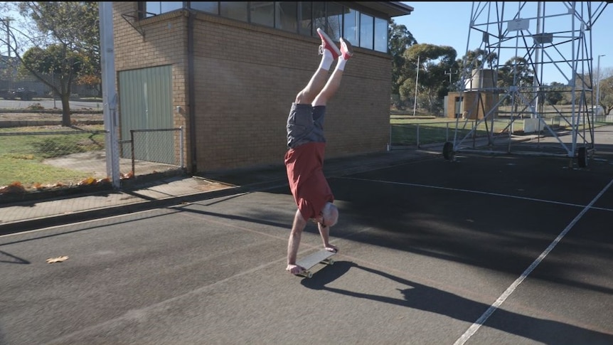 A man in a red shirt and grey shorts does a handstand on a skateboard on an asphalt surface 