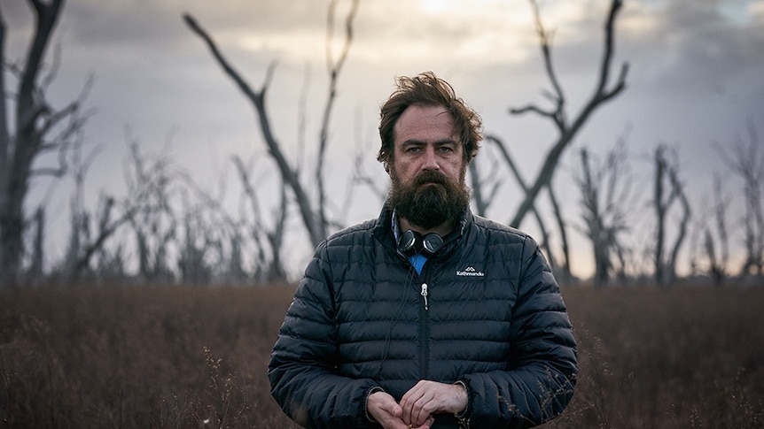 A man wearing puffer jacket and headphones around neck stands in outback desert landscape amongst dead trees on overcast day.