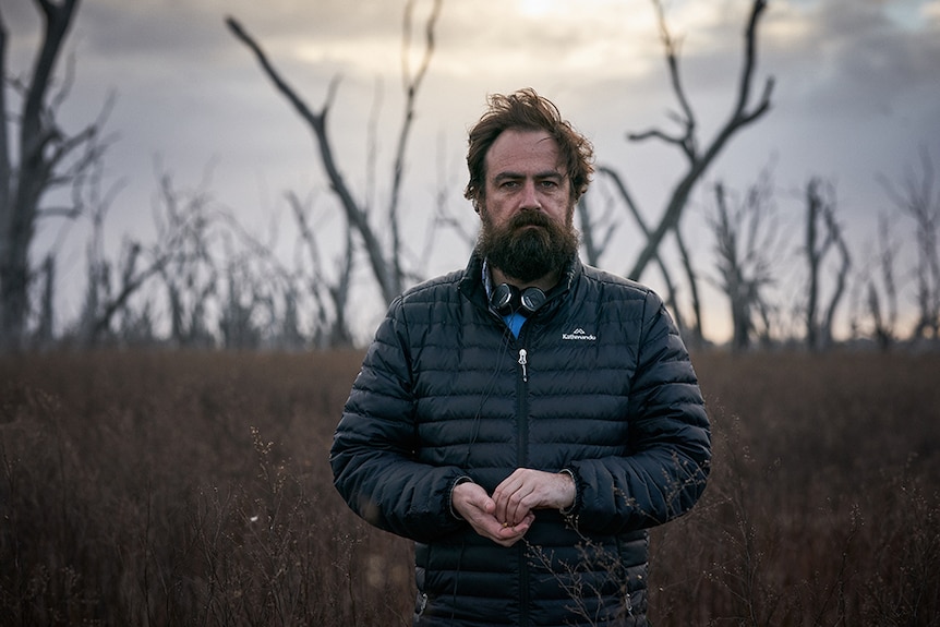 A man wearing puffer jacket and headphones around neck stands in outback desert landscape amongst dead trees on overcast day.