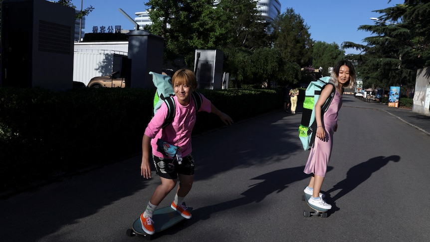 Two women dressed in pink with green backpacks on skate down a street. 