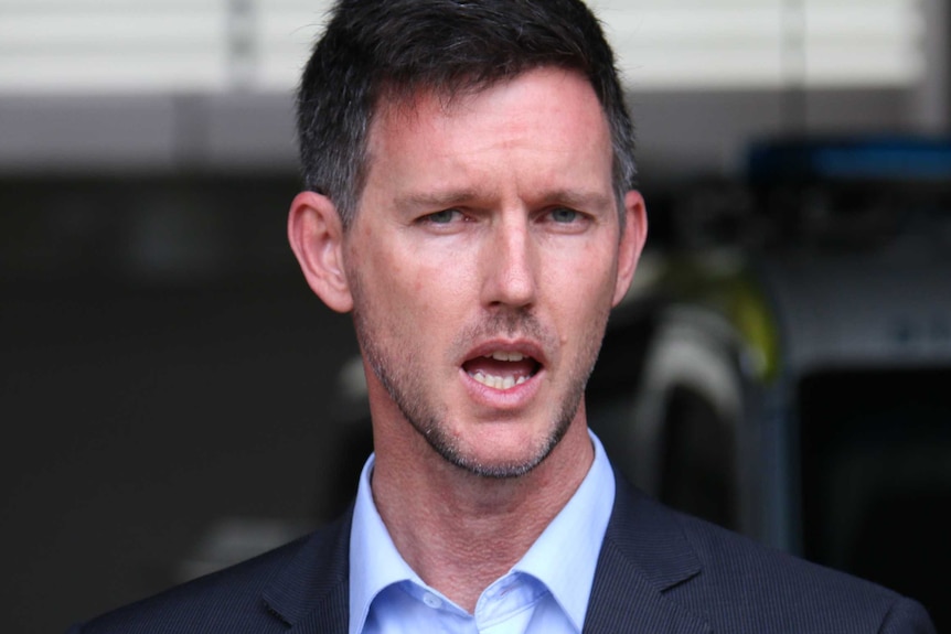 A man with short, dark hair and stubble, wearing a suit and speaking outside.