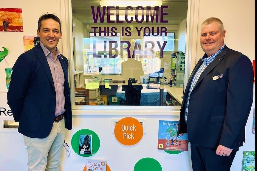 Two smiling men stand in a school building in front of a sign that says 'this is your library".