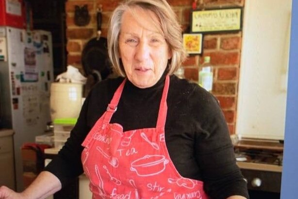 An elderly woman with grey hair, black top and pink apron smiles at the camera