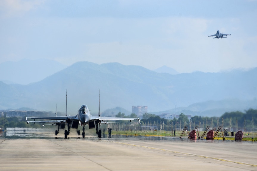 Jet militaire vu sur le tarmac alors qu'un autre est vu dans le ciel au-dessus.