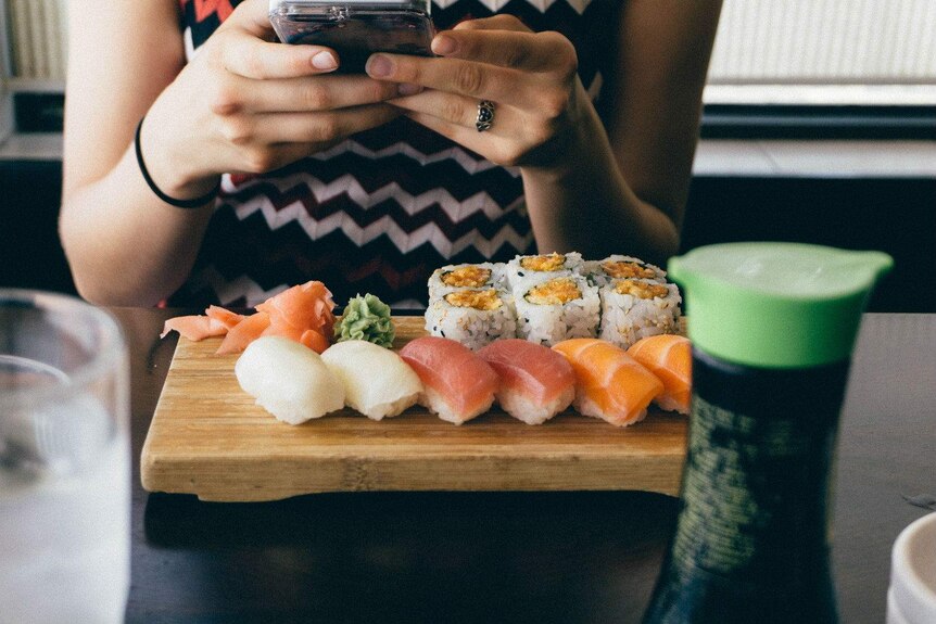 A woman with a phone sits down with a plate of sushi.