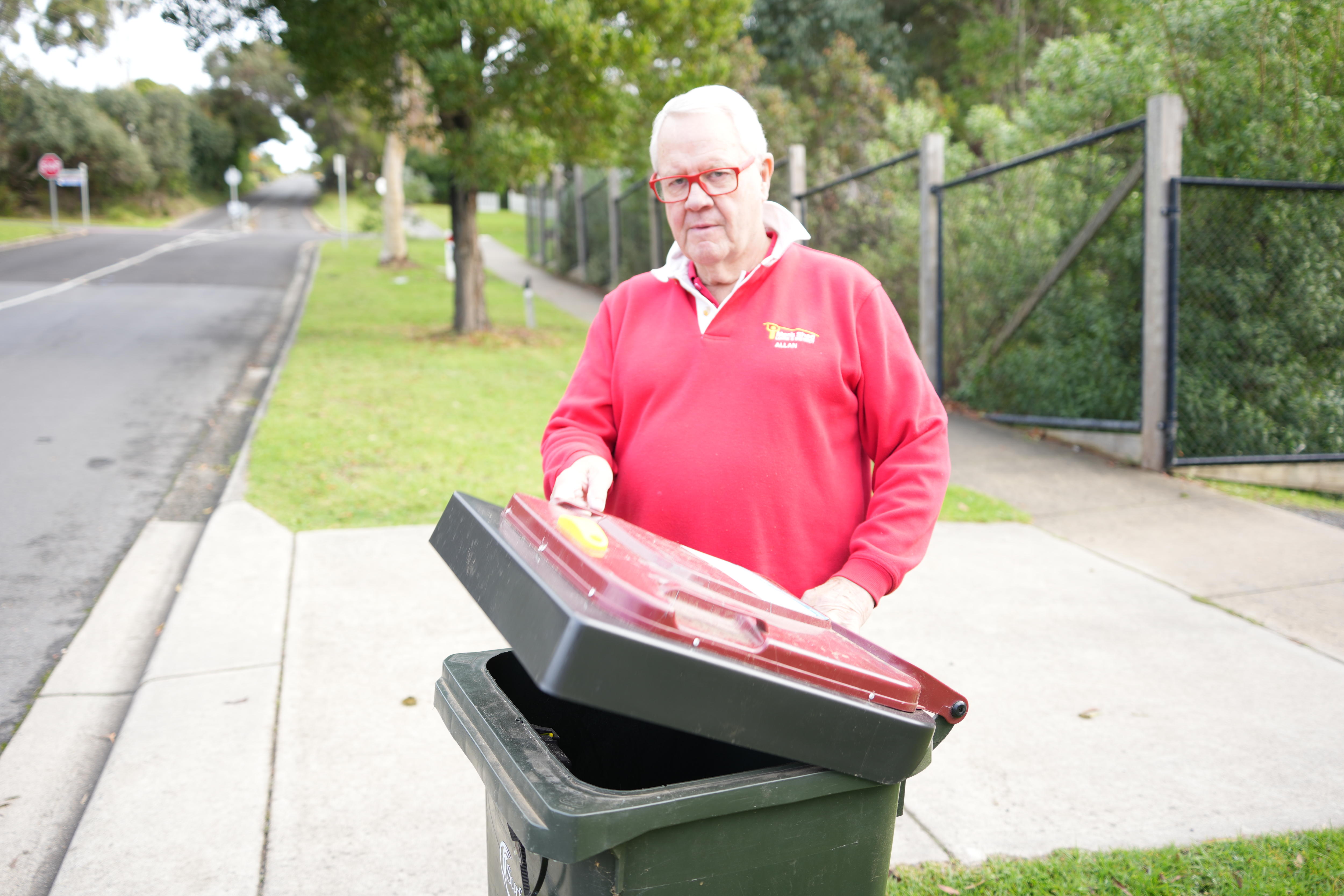A white haired man with red rimmed glasses and a red top holds open the lid of a wheelie bin in a street.