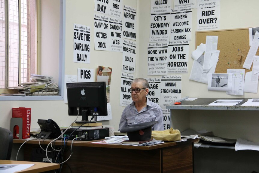 A man sits at in front of a computer screen at his work desk. Newspaper articles are hung up behind him