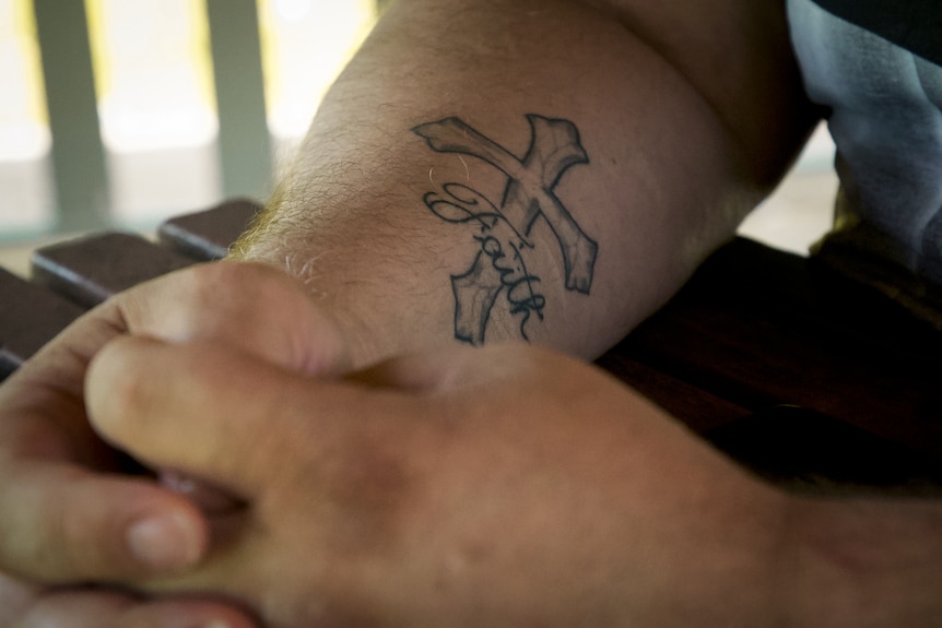 Close-up of a man's arm with a cross tattoo and the word 'faith'.