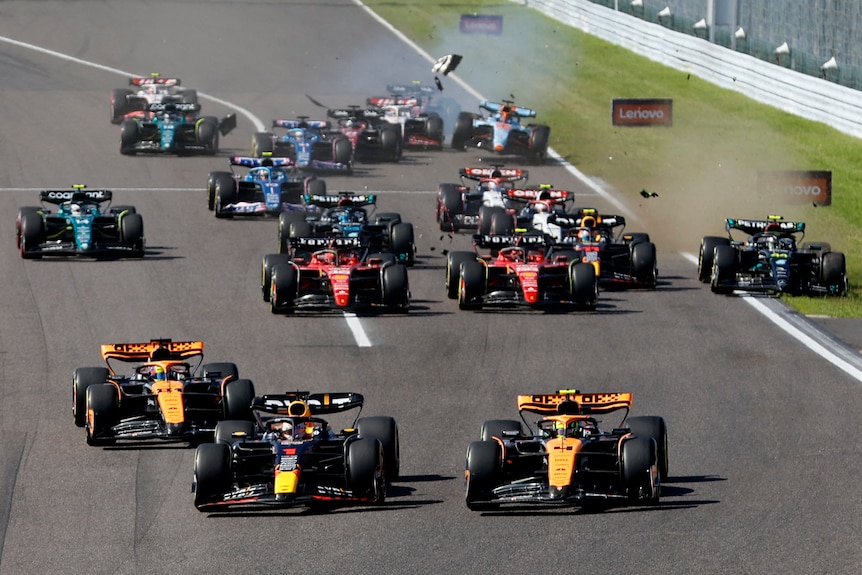 The full field of F1 cars racing to the first corner, with several colliding in the background