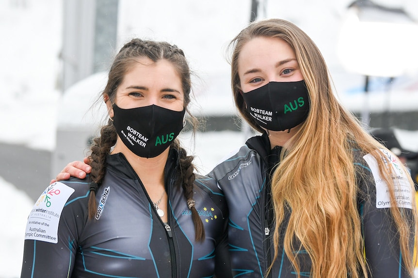 Bree Walker and Sarah Blizzard put their arms around each other and smile while wearing face masks.