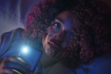 A young woman of colour with pink-dyed tight curls and blood on her face holds a phone with its light shining toward the camera