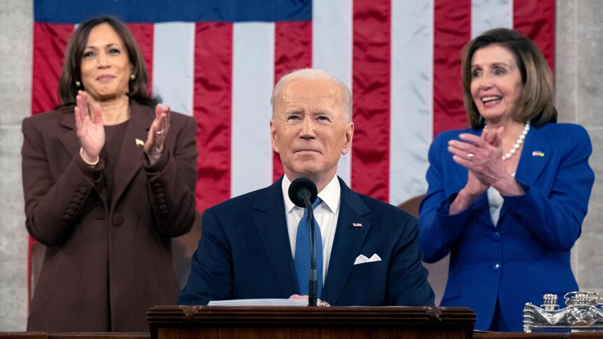 Joe Biden before a microphone while Kamala Harris and Nancy Pelosi applaud behind him