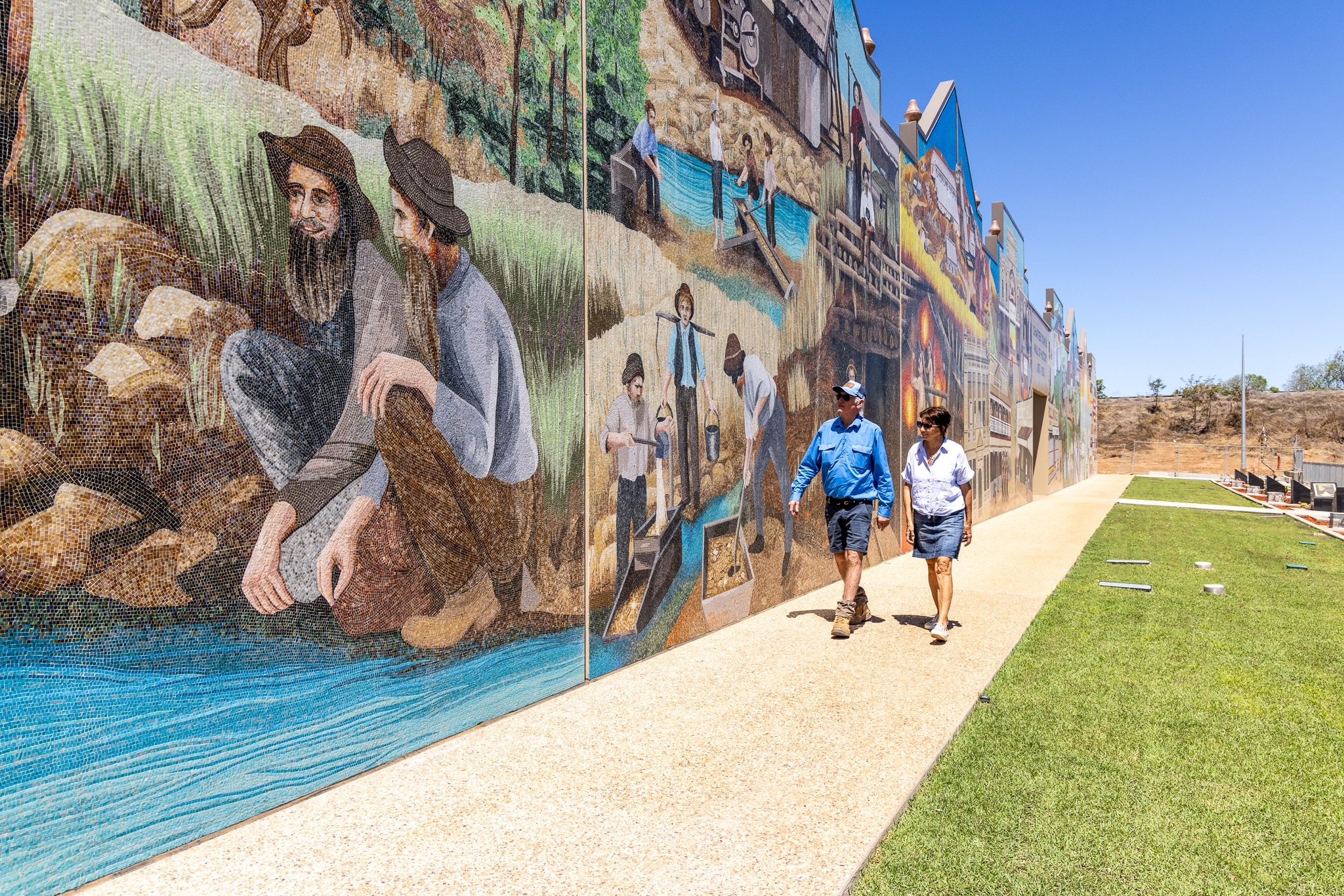 A man and woman walk past a large mosaic mural depicting historical scenes