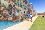 A man and woman walk past a large mosaic mural depicting historical scenes