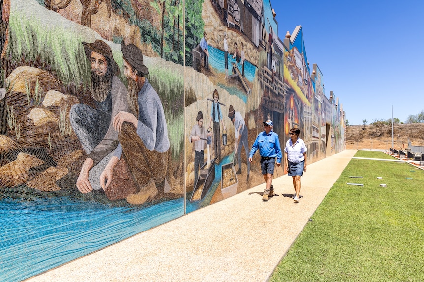 A man and woman walk past a large mosaic mural depicting historical scenes