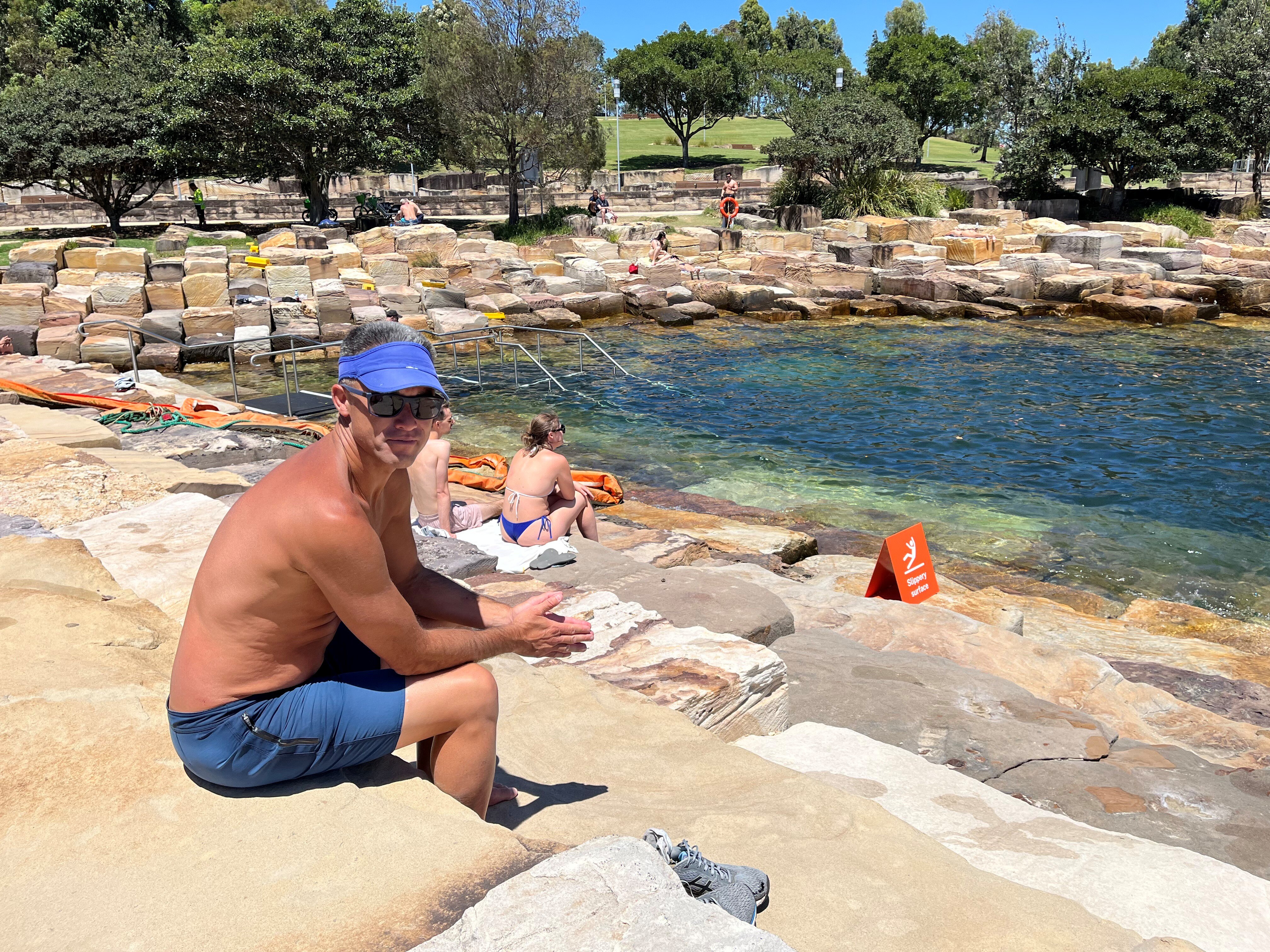 Mr Ching in blue swim shorts, a visor and sunglasses sits on a large slab of rock near the swimming area on a hot day.