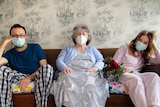 A man, an older lady and a woman all in masks sitting on a couch