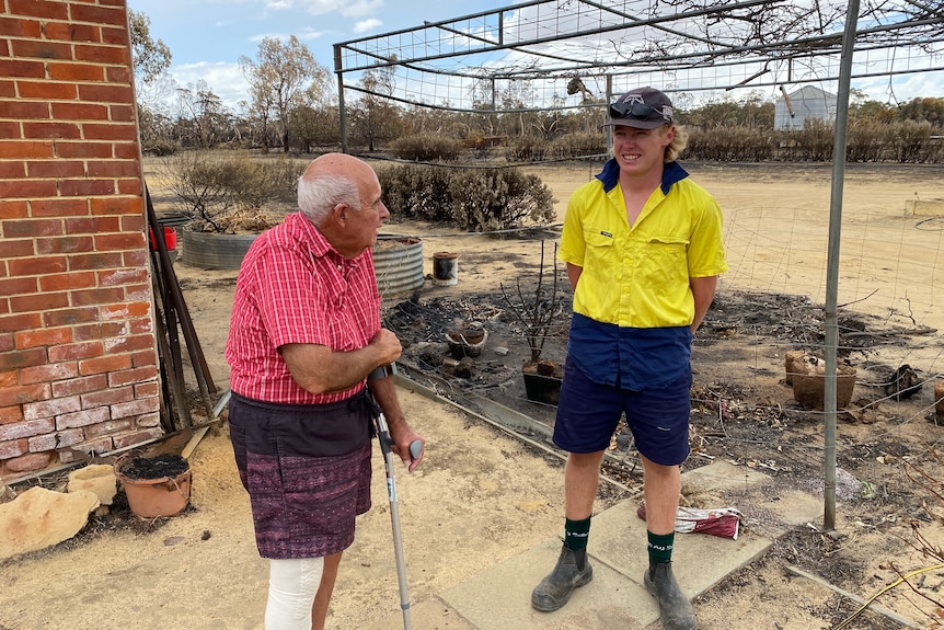 An elderly man and a younger man stand on a rural property blackened by bushfire