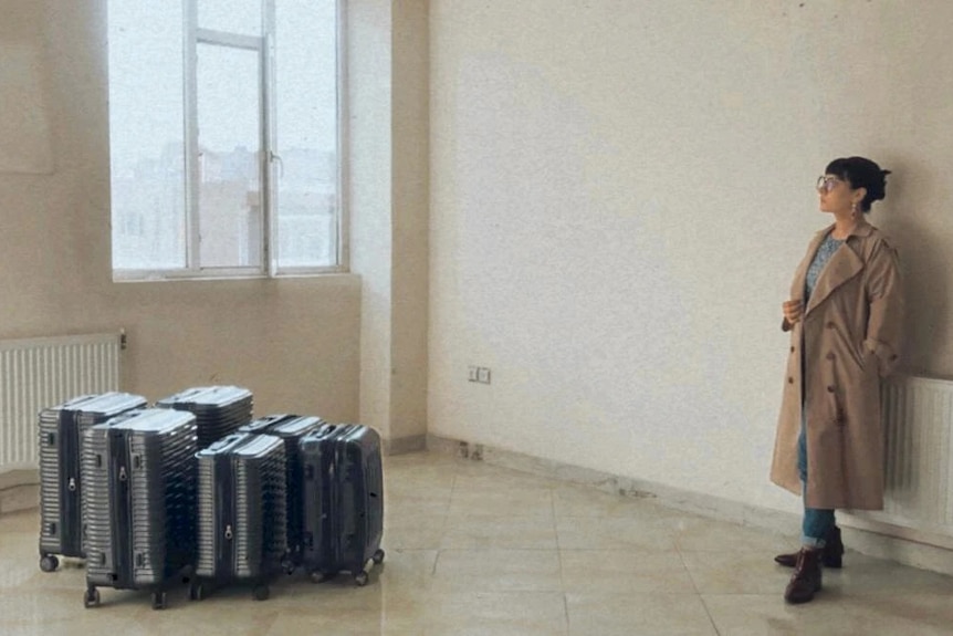 An Afghan woman in a trenchcoat stands in a bare room with a collection of suitcases at the centre