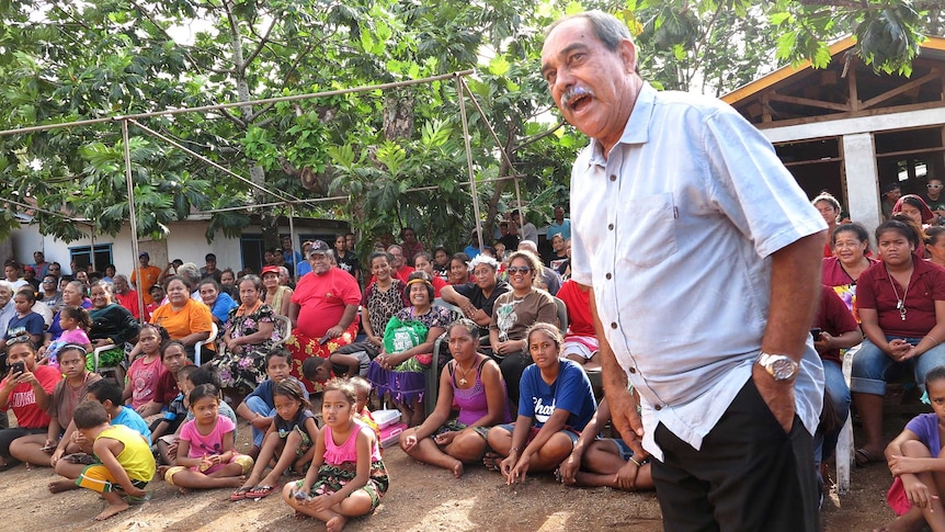 President Peter Christian stands in front of a large group of villagers in their village.