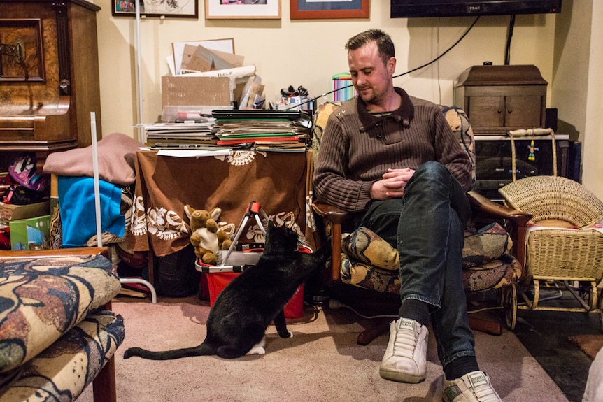 Nick Tonkin sitting on a armchair looking down at a black cat.