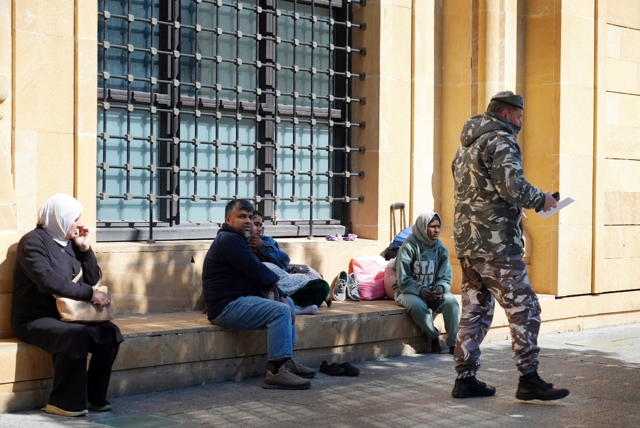 Un hombre con uniforme militar pasa junto a personas sentadas afuera en una escalera.
