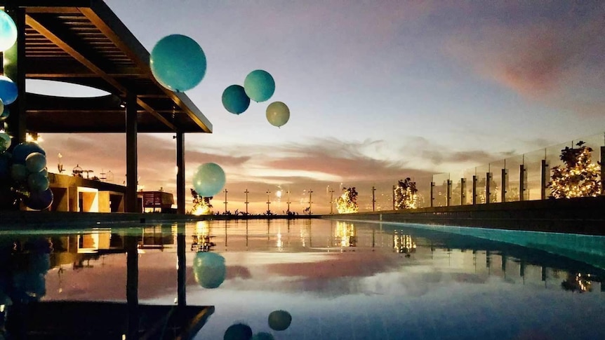 Large blue balloons hover over a rooftop pool with a setting sun in the background.