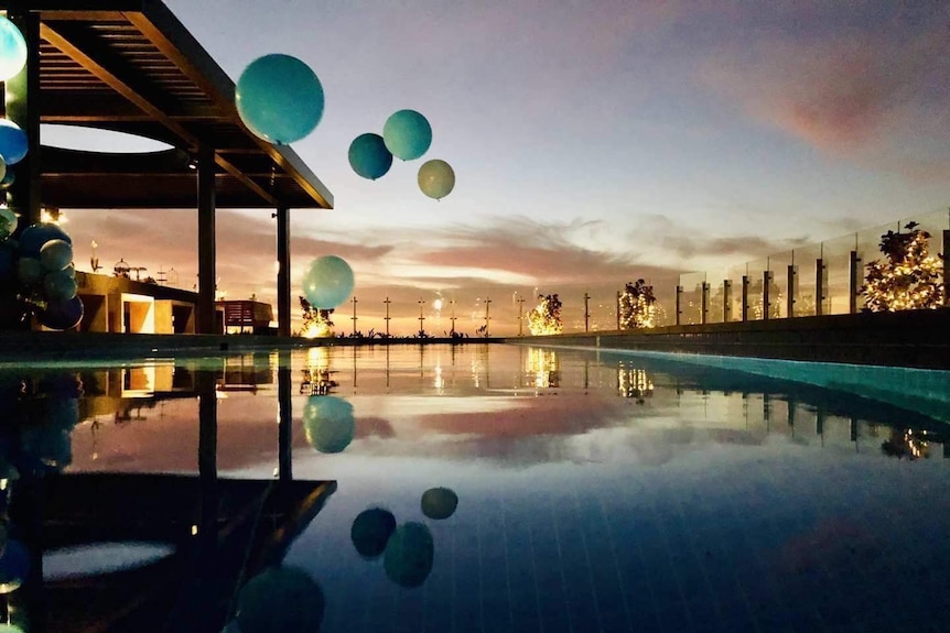 Large blue balloons hover over a rooftop pool with a setting sun in the background.