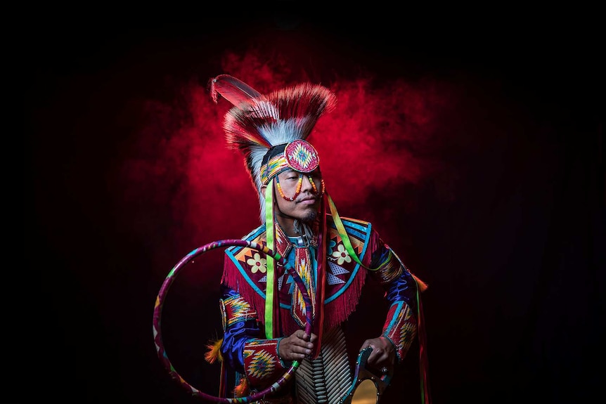 A man in bright coloured Native American regalia poses with hoop and mirror, stands in front of red smoke looking to his left.
