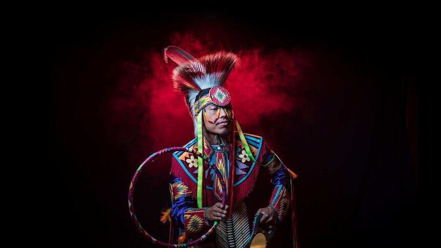 A man in bright coloured Native American regalia poses with hoop and mirror, stands in front of red smoke looking to his left.