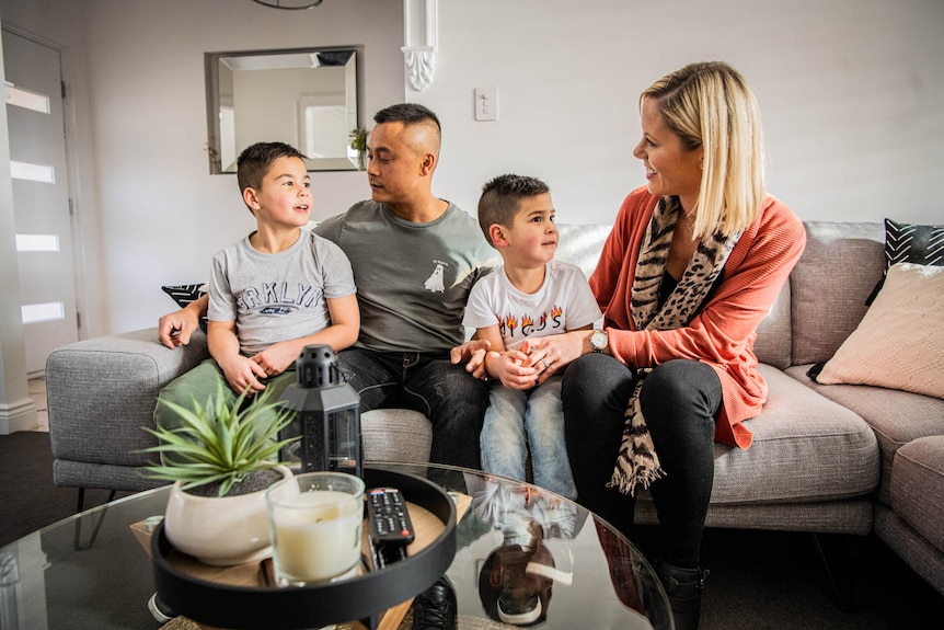 A young family sits together on a grey couch in a living room