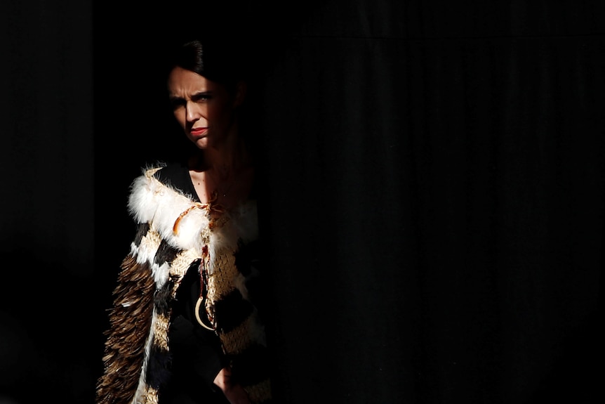 Jacinda Ardern, wearing a cloak, steps into a small sliver of light against a dark background