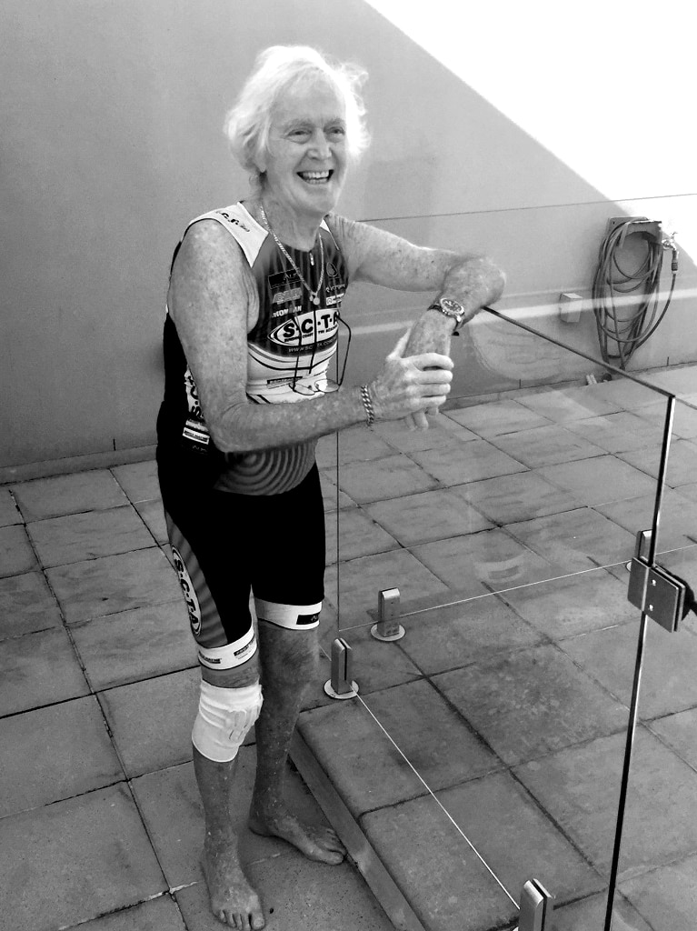 Man stands smiling not looking at the camera, wearing triathlon swimwear, leaning on a glass pool fence.