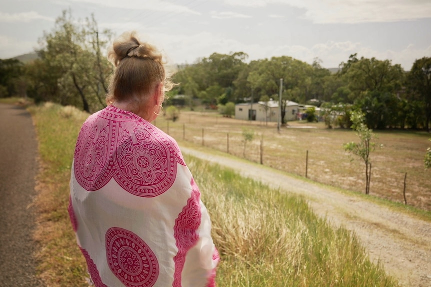 A woman is shown from behind looking over a fence into a property in a rural area.