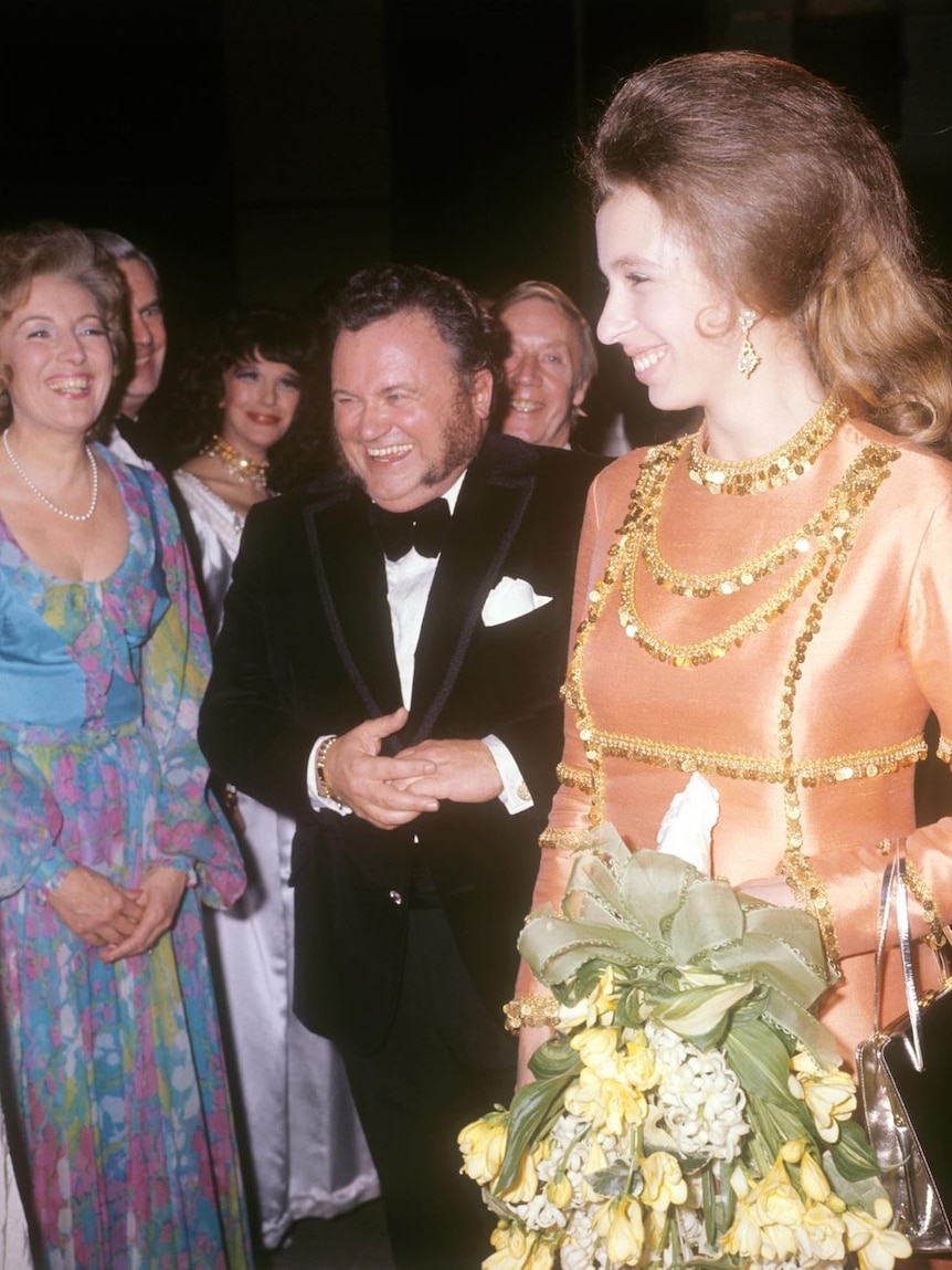 Princess Anne in a gold and orange dress holding a bouquet while surrounded by smiling people