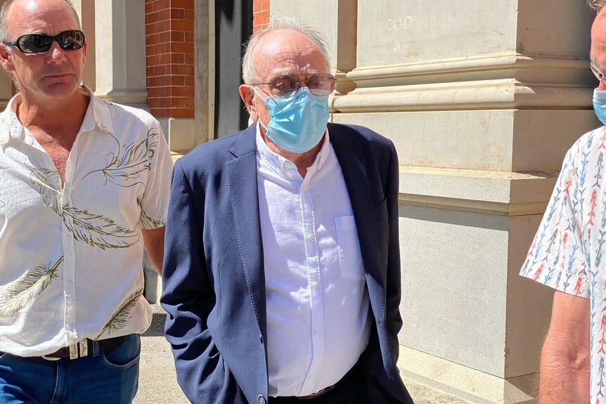 An old man wearing a surgical mask flanked by a man on each side walking outside court. 