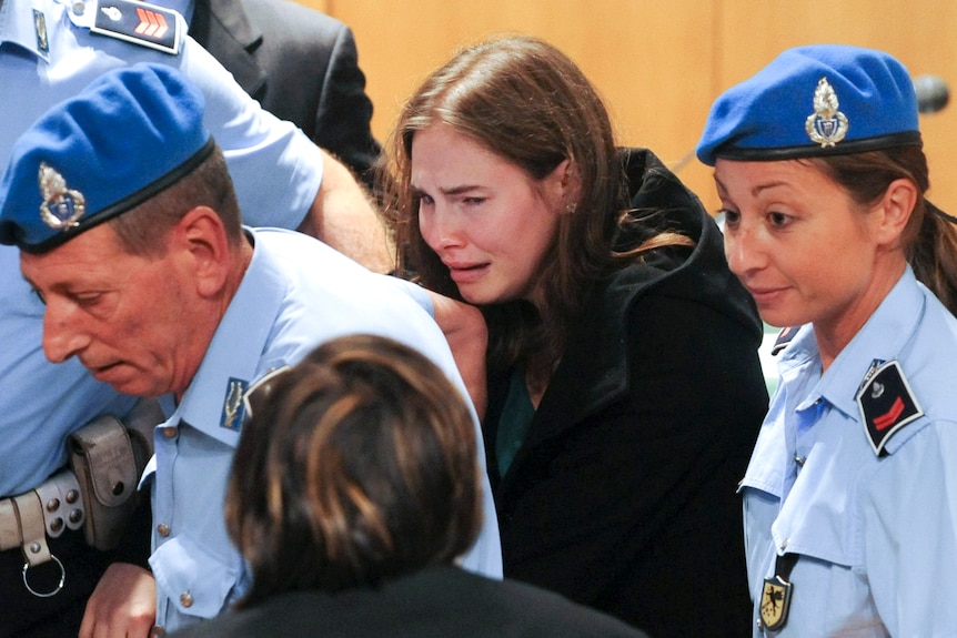 Two blue-uniformed prison officers flank Amanda Knox, with shoulder-length brown hair, who cries and looks distressed.