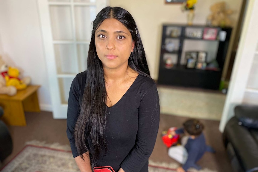 A woman with long black hair stands in her living room with her toddler playing on the floor behind her.