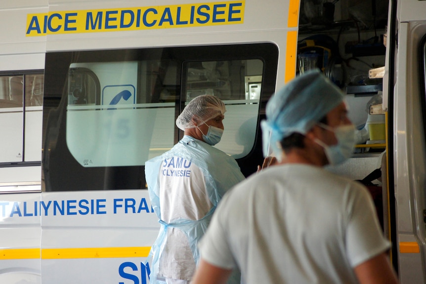 Несколько медицинских работников в средствах индивидуальной защиты стоят рядом с машиной скорой помощи