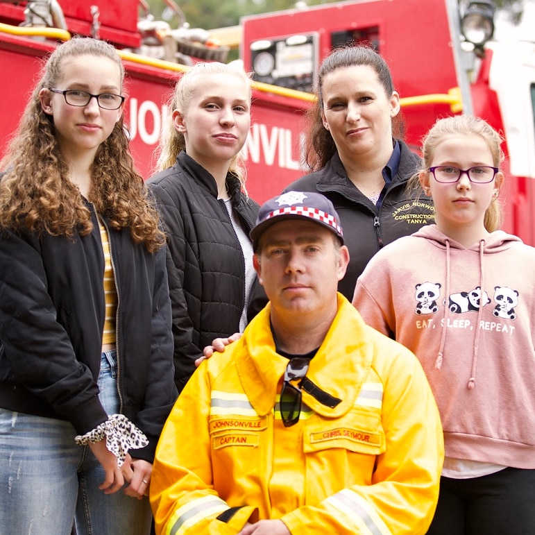 Chris Seymour kneels with his wife and three daughters behind him and his youngest daughter's hand on his shoulder.