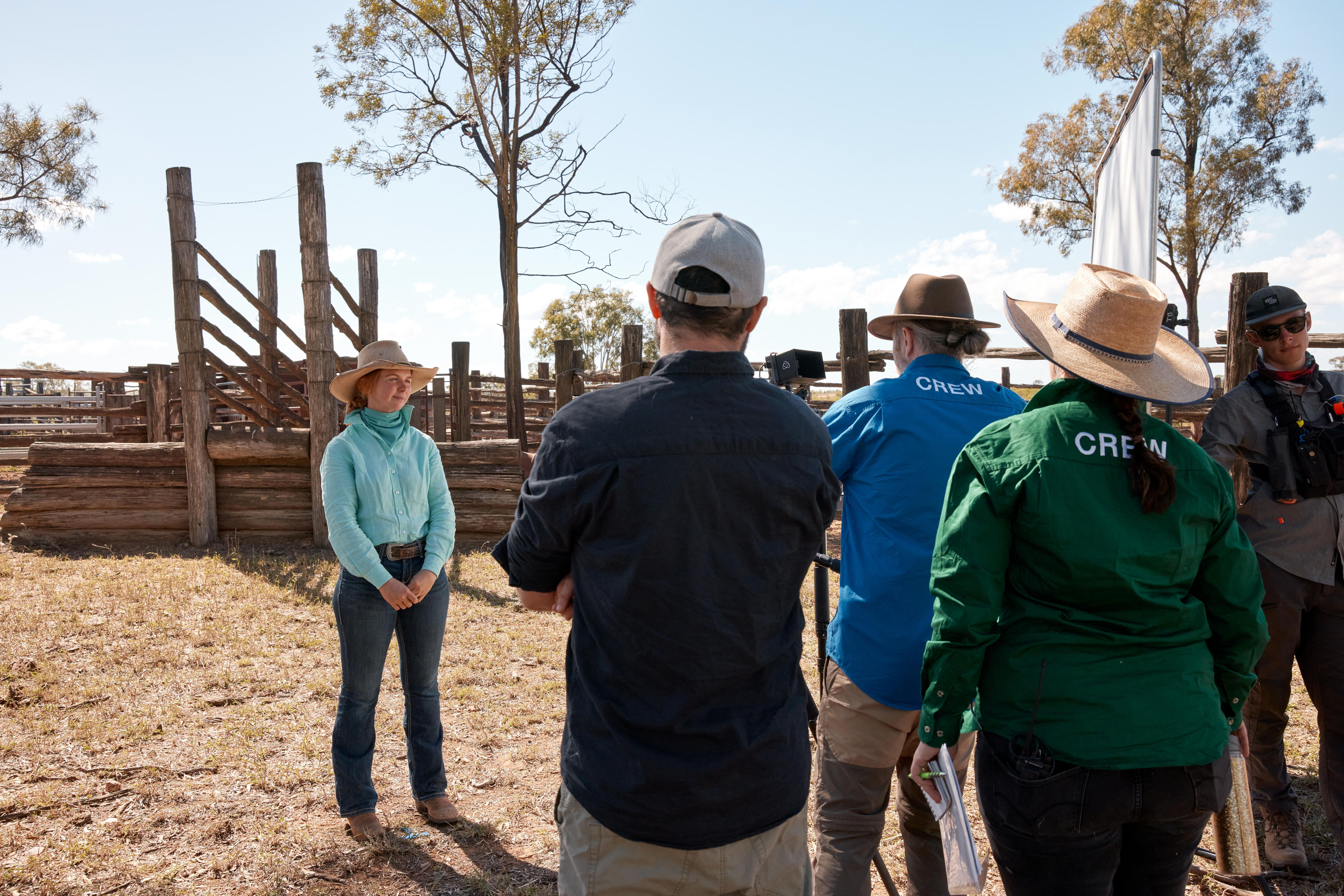 A camera crew in front of a woman in mustering gear standing near a cattleyard.