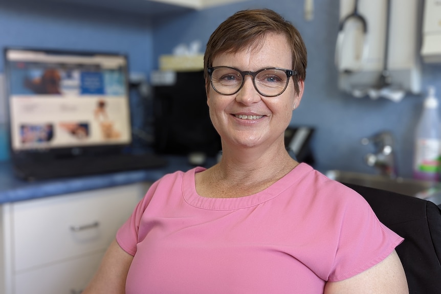A smiling woman in glasses and a pink t-shirt sits in a veterinary office 