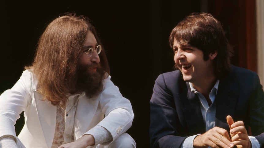 A bearded John Lennon in white and clean shaven Paul McCartney in navy talk and smile while seated