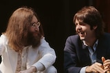 A bearded John Lennon in white and clean shaven Paul McCartney in navy talk and smile while seated